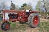 A vintage red Farmall model 656 tractor is parked on a patch of grass and dirt. The tractor shows signs of age, with some rust and fading paint. Large rear tires are prominent, and the engine components are partially visible. In the background, there are trees with budding leaves indicative of spring, and some logs are scattered nearby.