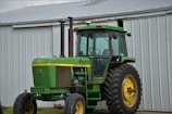 A green tractor with large yellow wheels is parked outside a metal barn. The tractor has a vertical exhaust stack and a cabin with glass windows. The barn is made of corrugated metal panels with a slanted roof.