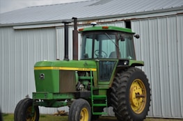 A green tractor with large yellow wheels is parked outside a metal barn. The tractor has a vertical exhaust stack and a cabin with glass windows. The barn is made of corrugated metal panels with a slanted roof.
