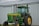 A green tractor with large yellow wheels is parked outside a metal barn. The tractor has a vertical exhaust stack and a cabin with glass windows. The barn is made of corrugated metal panels with a slanted roof.
