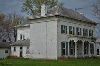 A large, old white brick house with green shutters and an ornate porch sits surrounded by grassy lawns. The building appears weathered, with visible signs of paint peeling. In the background, there are bare trees, hinting at the season being late winter or early spring.