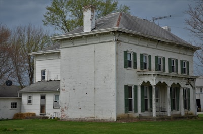 A large, old white brick house with green shutters and an ornate porch sits surrounded by grassy lawns. The building appears weathered, with visible signs of paint peeling. In the background, there are bare trees, hinting at the season being late winter or early spring.