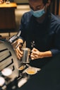 A barista wearing a face mask is preparing coffee at an espresso machine. They are holding a metal pitcher and pouring milk into a coffee cup on the counter.