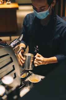 A barista wearing a face mask is preparing coffee at an espresso machine. They are holding a metal pitcher and pouring milk into a coffee cup on the counter.