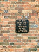 A brick wall with a sign that reads: 'On this very spot in 1649 nothing at all happened.' The sign is made of a dark material with light-colored lettering, creating a humorous and ironic message.