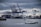 Container ships docked at a bustling port with multiple cranes loading and unloading cargo. A smaller patrol boat sails by in the foreground under a cloudy sky.