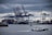 Container ships docked at a bustling port with multiple cranes loading and unloading cargo. A smaller patrol boat sails by in the foreground under a cloudy sky.