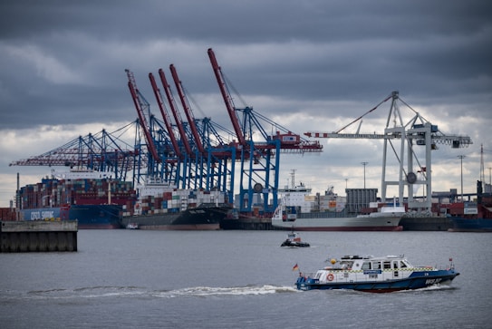Container ships docked at a bustling port with multiple cranes loading and unloading cargo. A smaller patrol boat sails by in the foreground under a cloudy sky.