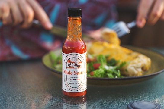 A bottle of Mule Sauce is prominently placed on a table with a meal in the background. The bottle features a label with an image of a mule and text. In the blurred background, someone is holding a fork and knife, possibly cutting into a dish that includes leafy greens and a portion of food, perhaps a burrito or similar item.