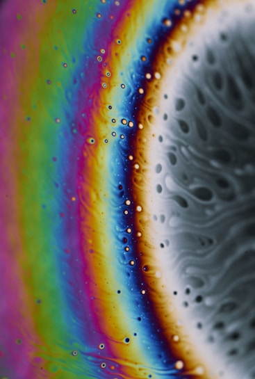 Vibrant, swirling patterns of colors, including blues, greens, and purples, transitioning smoothly into oranges and yellows. The texture appears fluid and organic, resembling soap film or oil on water, with tiny bubbles dispersed throughout.