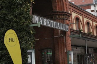 An urban scene featuring a brick building with a sign reading 'Markthalle'. The building has arch windows and is adorned with greenery. In the foreground, there is a yellow flag with the word 'LIFE' visible. The structure seems to be part of a public market or similar venue.