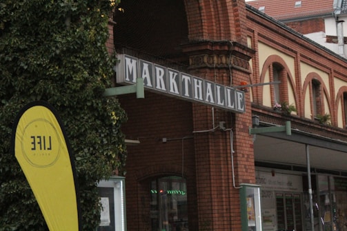An urban scene featuring a brick building with a sign reading 'Markthalle'. The building has arch windows and is adorned with greenery. In the foreground, there is a yellow flag with the word 'LIFE' visible. The structure seems to be part of a public market or similar venue.