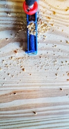 A close-up of a woodworking scene featuring a blue Kreg pocket hole jig partially covered with wood shavings. The jig is placed on a light-colored wooden board with visible grain patterns. A drill with a red handle is inserted into the jig, ready to make precise holes.