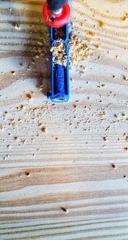 A close-up of a woodworking scene featuring a blue Kreg pocket hole jig partially covered with wood shavings. The jig is placed on a light-colored wooden board with visible grain patterns. A drill with a red handle is inserted into the jig, ready to make precise holes.