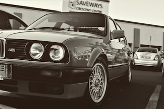 A classic car parked in a lot, featuring unmistakable round headlights and a distinctive grille. The wheels have intricate designs. In the background, a building with signage reads 'SAVEWAYS CRESCENT CENTRE', and another car is visible.