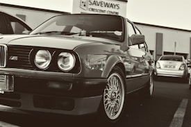 A classic car parked in a lot, featuring unmistakable round headlights and a distinctive grille. The wheels have intricate designs. In the background, a building with signage reads 'SAVEWAYS CRESCENT CENTRE', and another car is visible.