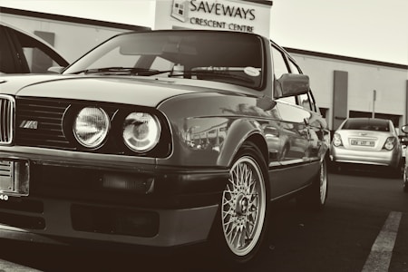 A classic car parked in a lot, featuring unmistakable round headlights and a distinctive grille. The wheels have intricate designs. In the background, a building with signage reads 'SAVEWAYS CRESCENT CENTRE', and another car is visible.