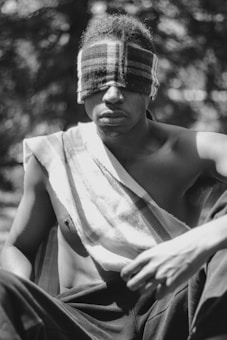 A person sitting outdoors with a plaid fabric wrapped around as a makeshift blindfold. Sunlight filters through trees in the background, creating a dappled pattern on the person's bare upper body. They appear to be deep in thought or meditation.