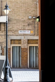The image captures a narrow alley with a tall, vintage-style street lamp on the left. The walls are made of light brown bricks, featuring a street sign that reads 'Vine Street W1, City of Westminster'. Two closed metal doors are set into the wall, framed by faded bricks and a concrete sidewalk leading into the alley. Electrical wires and a security light are also visible on the wall.
