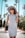A fashionable woman wearing a fitted grey dress and a large sun hat holds a handbag while walking down a palm-lined street. She is also wearing sunglasses, exuding a chic and confident style.