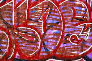An abstract graffiti artwork featuring bold, rounded shapes filled with red and brown lines against a purple background. White outlines add contrast and definition to the overlapping forms.
