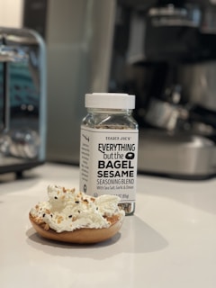 A jar of 'Everything but the Bagel Sesame Seasoning Blend' sits on a white countertop next to a bagel half topped with cream cheese and sprinkled with the seasoning. The background includes blurred kitchen appliances, indicating a kitchen setting.