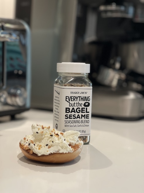 A jar of 'Everything but the Bagel Sesame Seasoning Blend' sits on a white countertop next to a bagel half topped with cream cheese and sprinkled with the seasoning. The background includes blurred kitchen appliances, indicating a kitchen setting.