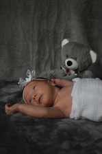 A newborn baby lies on a soft, dark textured surface, wrapped in a white cloth. The infant has a white headband with a bow and gazes upwards. Beside the baby, a plush gray teddy bear sits in the background.