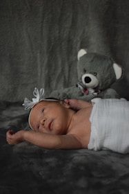 A newborn baby lies on a soft, dark textured surface, wrapped in a white cloth. The infant has a white headband with a bow and gazes upwards. Beside the baby, a plush gray teddy bear sits in the background.