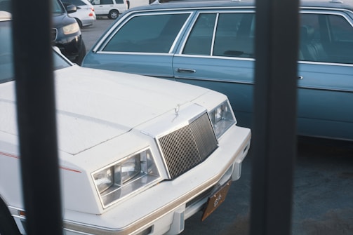 Two parked cars are seen, one white and one blue, captured through metal bars. The white car is at the forefront, displaying a classic grille and square headlights, while the blue car is partially visible in the background.