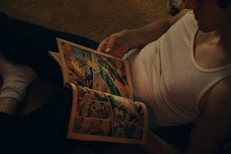 A person is sitting on the floor wearing a sleeveless shirt and holding an open comic book. The setting has a dim, warm lighting, giving a cozy and relaxed atmosphere.
