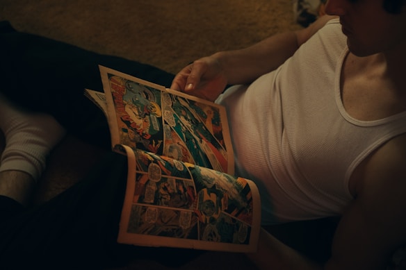 A person is sitting on the floor wearing a sleeveless shirt and holding an open comic book. The setting has a dim, warm lighting, giving a cozy and relaxed atmosphere.