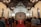 A congregation gathers inside a chapel with a wooden ceiling and chandeliers. People stand in rows facing the altar, adorned with a cross and flickering candles. Musicians are positioned to the right, surrounded by a warm, inviting atmosphere.