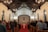 A congregation gathers inside a chapel with a wooden ceiling and chandeliers. People stand in rows facing the altar, adorned with a cross and flickering candles. Musicians are positioned to the right, surrounded by a warm, inviting atmosphere.
