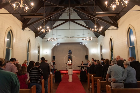 A congregation gathers inside a chapel with a wooden ceiling and chandeliers. People stand in rows facing the altar, adorned with a cross and flickering candles. Musicians are positioned to the right, surrounded by a warm, inviting atmosphere.