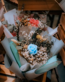 A bouquet of flowers featuring a striking blue rose at the center, surrounded by small white flowers and various dried elements. The arrangement has a rustic and artisanal feel, enhanced by soft pastel paper wrapping.