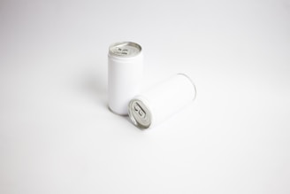 Two plain aluminum beverage cans with a white label are positioned on a smooth, white surface. One can is upright while the other lies horizontally beside it.