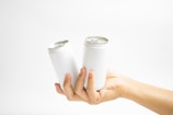 A hand holds two white aluminum cans against a plain white background. The cans are plain with no labels, and they are positioned upright with the opening tabs visible. The lighting is bright and even, emphasizing the minimalist aesthetic.
