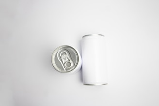 A pair of aluminum cans are placed on a plain white background. One can is viewed from the top, showcasing the pull-tab, while the other is lying on its side, displaying its cylindrical shape.