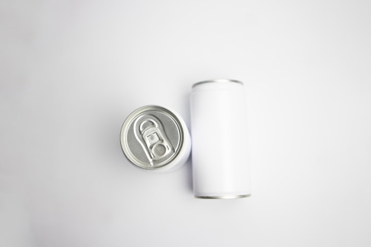 A pair of aluminum cans are placed on a plain white background. One can is viewed from the top, showcasing the pull-tab, while the other is lying on its side, displaying its cylindrical shape.
