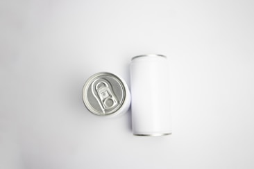 A pair of aluminum cans are placed on a plain white background. One can is viewed from the top, showcasing the pull-tab, while the other is lying on its side, displaying its cylindrical shape.