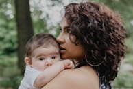 A woman with curly dark hair is holding a baby close to her shoulder in a natural, outdoor setting. The baby, dressed in a white outfit, looks curiously at the camera, while the woman gazes thoughtfully into the distance. The background is filled with greenery, suggesting a forest or park environment.