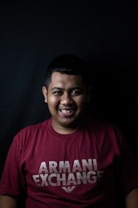 A person is smiling while wearing a maroon t-shirt with 'Armani Exchange' written on it. The background is a dark solid color, enhancing the subject's presence.
