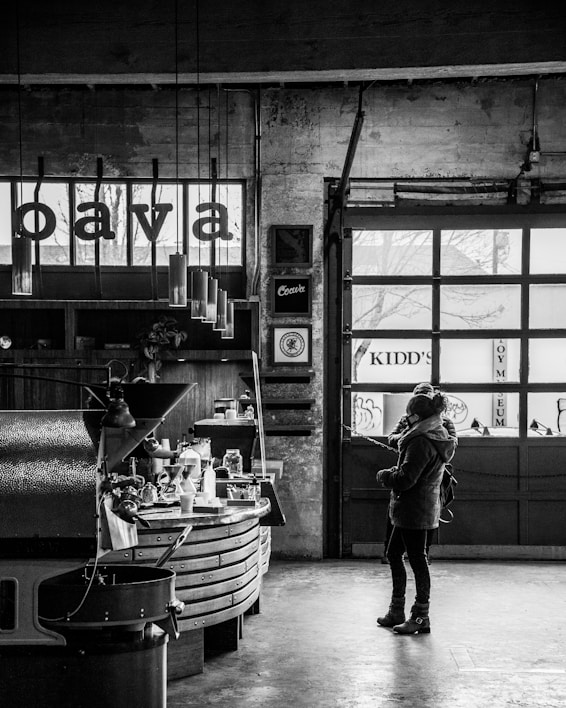 A person stands in an industrial-style coffee shop, examining a selection of coffee-making equipment on a counter. The setting is rustic and vintage with a raw, unfinished stone wall and large windows that let in natural light. Hanging pendant lights and various coffee gadgets are visible, enhancing the coziness of the space.