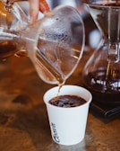 A freshly brewed coffee is being poured from a glass coffee maker into a white paper cup. The liquid appears dark and rich, with steam rising and condensation visible on the glass. The setting has a rustic ambiance with a wooden surface.