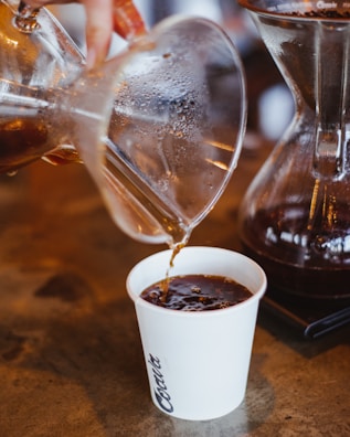 A freshly brewed coffee is being poured from a glass coffee maker into a white paper cup. The liquid appears dark and rich, with steam rising and condensation visible on the glass. The setting has a rustic ambiance with a wooden surface.