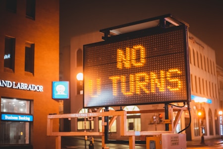A digital traffic sign is prominently displayed with the words 'NO TURNS' illuminated in bright orange. The background includes a few buildings, one of which has a sign indicating it might be a bank. It's nighttime, as suggested by the artificial lighting and the dark sky.