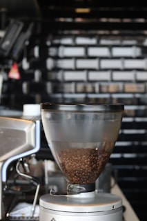 A coffee grinder filled with dark coffee beans is positioned in a modern kitchen setting. The background features a glossy black tile wall that adds a sleek and contemporary feel to the environment. The metallic components of the grinder reflect the light, highlighting its clean and polished design.