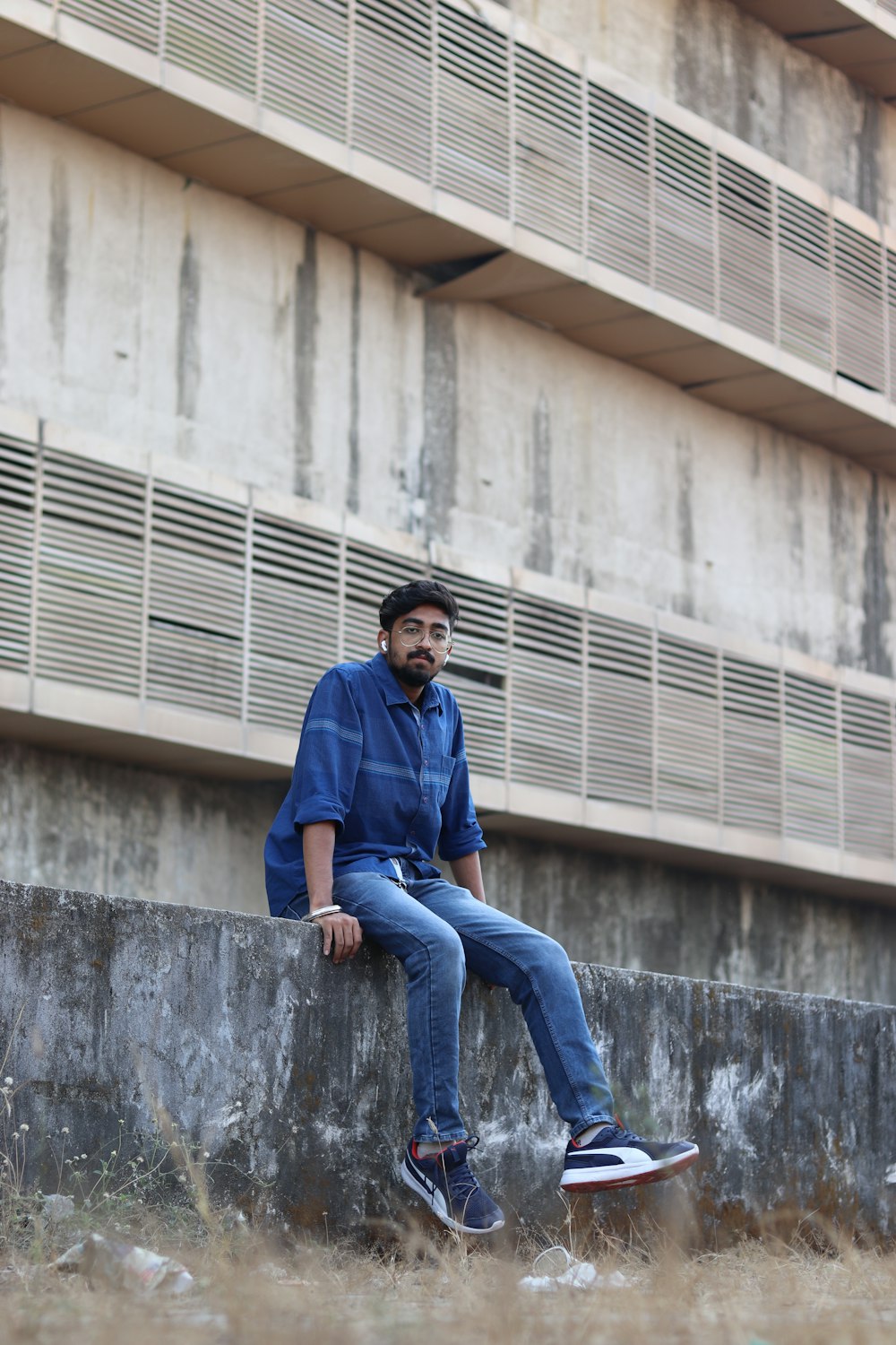 man in blue dress shirt and blue denim jeans sitting on concrete wall during daytime