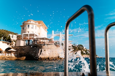 A picturesque view of a coastal area featuring a large, elegant villa perched atop a rocky cliff. The villa is surrounded by lush greenery and overlooks a sparkling body of water. Splashes of water are captured in motion against a clear blue sky. In the foreground, metal railings are visible, possibly indicating a seaside promenade or dock.